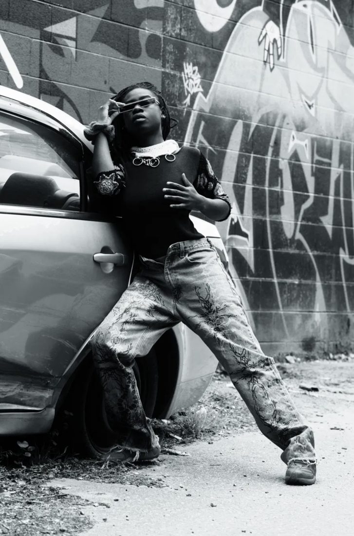 Black and white editorial photograph, figure leaning against a car by a graffiti wall
